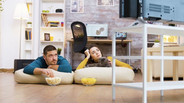 Young couple watching tv with their cat sitting on pillows for the floor while eating chips and popcorn.