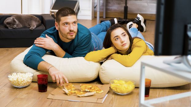 Attractive couple relaxing on pillows for the floor watching tv and eating pizza with their cat sleeping in the background.