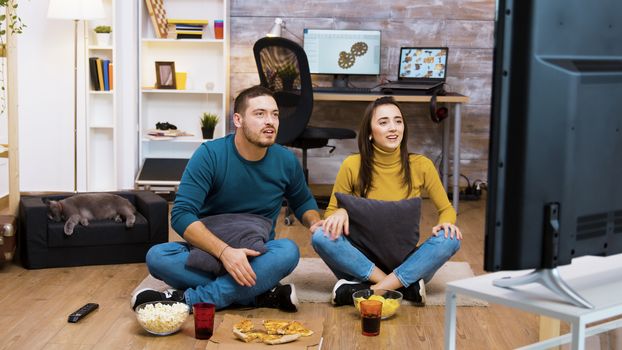 Caucasian young couple sitting on the floor in living room cheering up for their favorite sport team. Cat sleeping in the background.