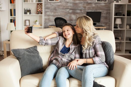 Mother and daughter looking into the phone camera and taking a selfie sitting on the couch in their apartment.