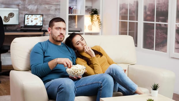 Caucasian couple relaxing watching tv in living room eating popcorn and chips.