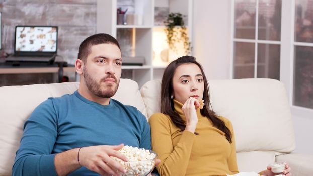Couple sitting on couch eating fried chicken and popcorn while watching tv. Scared couple after a scary moment in the movie.
