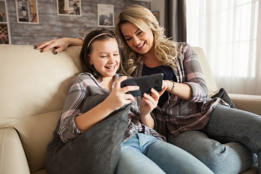 Happy young mother and her child smiling while using smartphone sitting on the couch.