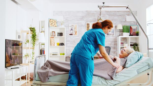 Female nurse helping an old lady to go to bed and covers her with a blanket in a nursing retirement home with big windows