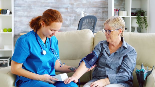 Female doctor taking blood pressure of senior woman in nursing home sitting on sofa