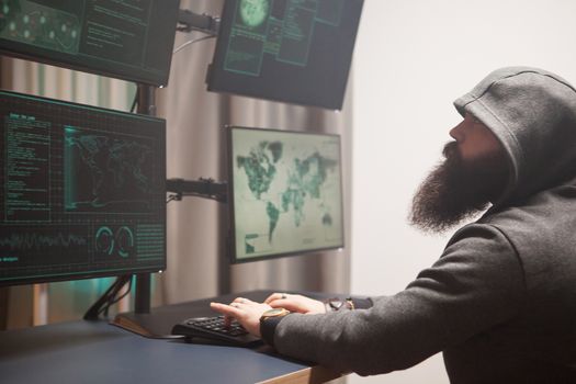 Side view of bearded male hacker wearing a hoodie launching a cyber attack in room with multiple screens.