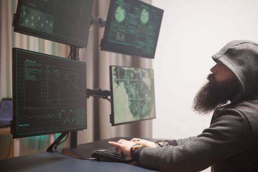 Bearded cyberterrorist wearing a hoodie writing a dangerous virus. Hacker using computer with multiple screens.