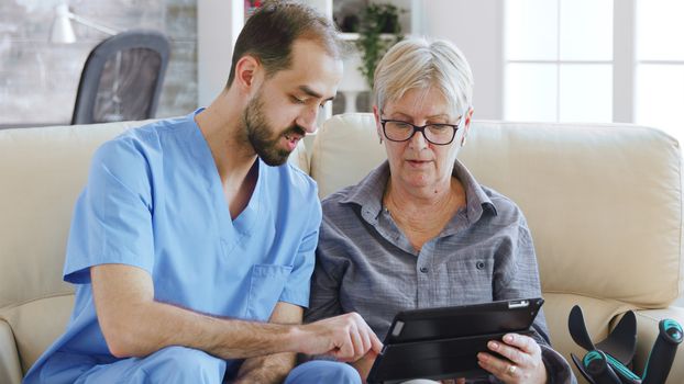 Male assistant teaching senior retired elderly old woman how to use her tablet computer in nursing home for her therapy