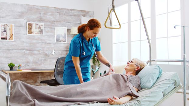 Female nurse helping an old disabled and retired woman to go to bed. The caregiver is covering her with a blanket.