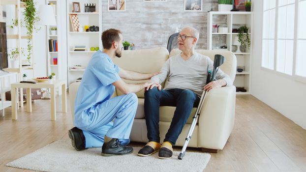 Revealing shot of male nurse checking on retired old man with alzheimer sitting on a couch in the nursing home