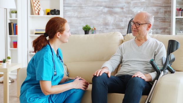 Female nurse talking and taking care of old disabled man sitting on the sofa in nursing home. Social worked and caregiver