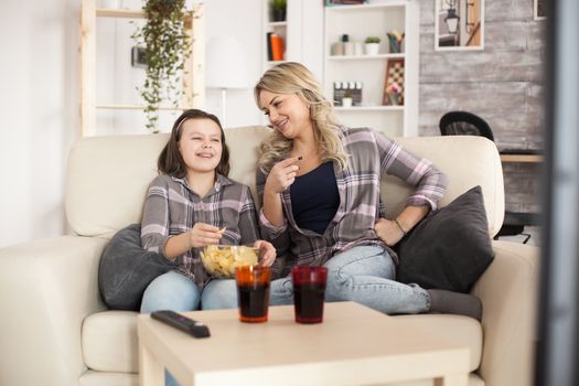 Little girl and her mother laughing while watching tv in living room eating chips and drinking soda.