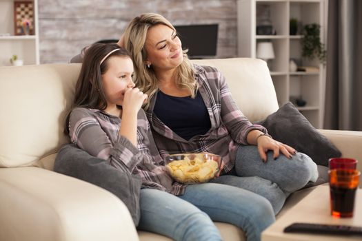 Cheerful daughter and her young mother relaxing watching a film on tv. Snacks and soda.