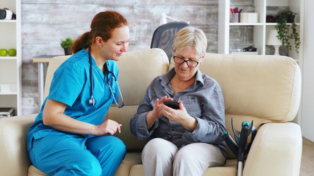 Female doctor in nursing home sitting on the couch teaching senior woman to use smartphone