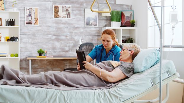 Female caregiver helps an old disabled woman lying in hospital bed to use a digital tablet PC. Bright room with big windows