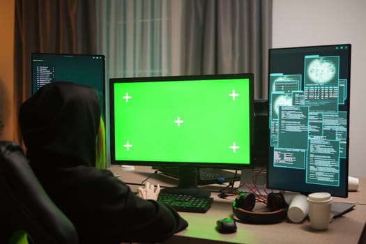 Dangerous hooded female hacker in front of computer with green screen.