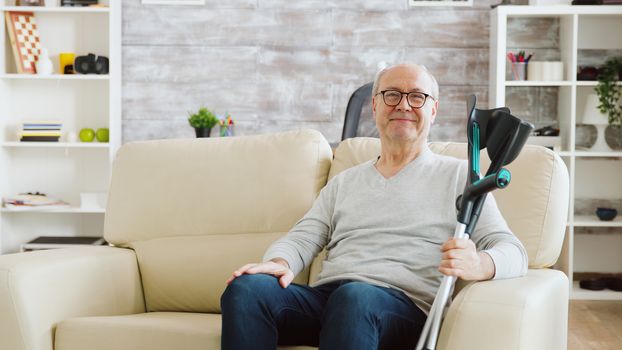 Portrait of elderly retired man smiling to the camera in cozy nursing home, he has crutches next to the sofa