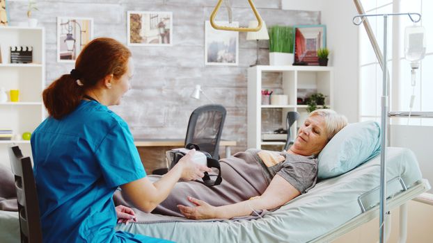 Female nurse in retirement home giving an old disabled lady a VR headset to ease the pain and distract her while she is lying in hospital bed