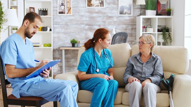 Male assistant taking notes on clipboard while female doctor is talking with senior woman in nursing home