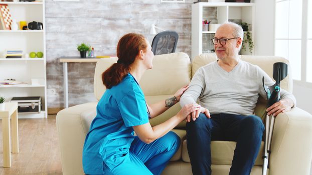 Female nurse looking after an old patient with alzheimer dissease sitting on the sofa in bright nursing home with big windows