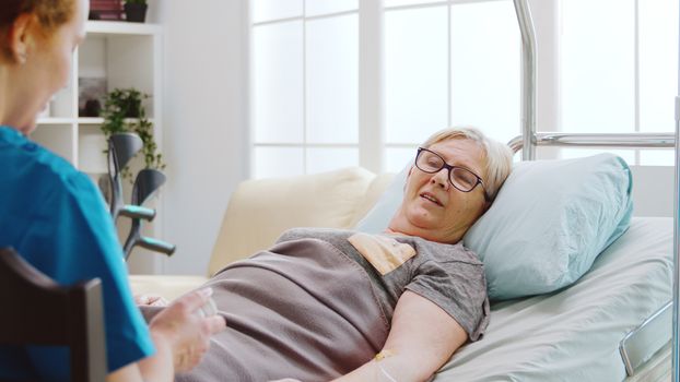 Nurse showing to an old lady what pills dossage to take while she is lying in hospital bed in retirement home