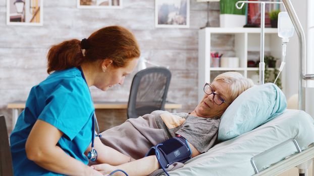 In nursing home old lady gets her blood pressure checked by an young caucasian nurse. Bright room with big windows