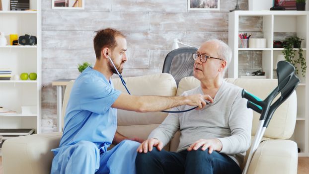 Revealing shot of young male nurse listening to old retired man heartbeat in a bright and cozy nursing home. Caregiver and social worker