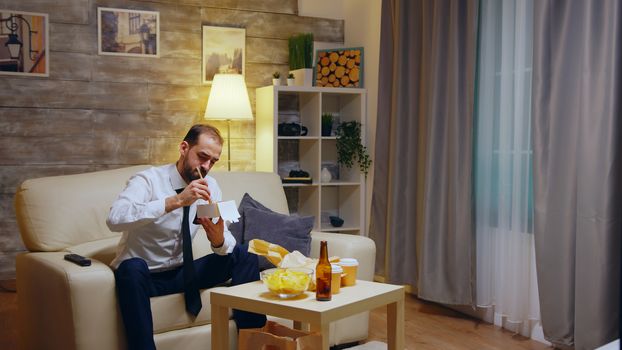 Businessman sitting on couch eating takeaway noodles from box after work in his apartment watching tv.