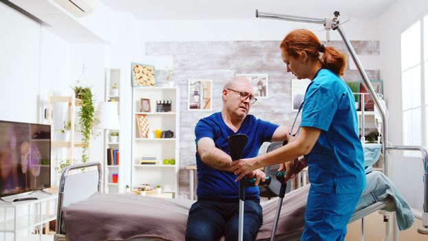 Nurse helping a disabled retired old man to stand up from his hospital bed in a bright and cozy nursing retirement home