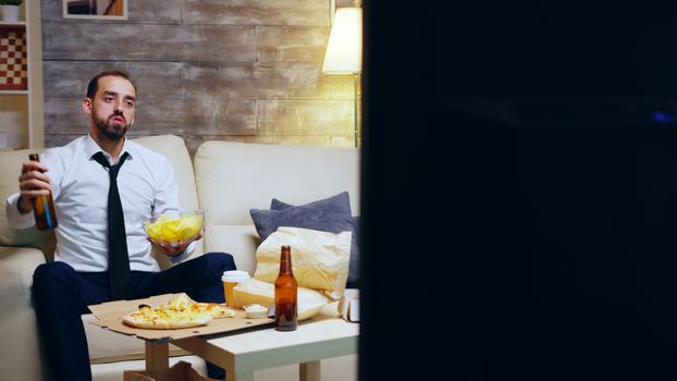Businessman still in his suit sitting on couch cheering while watching sports on tv. Entrepreneur holding chips.