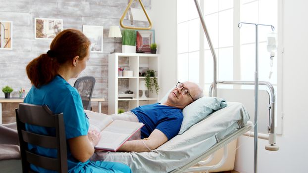 Old retired sick man lying in hospital bed in a nursing home while a nurse is reading him a book