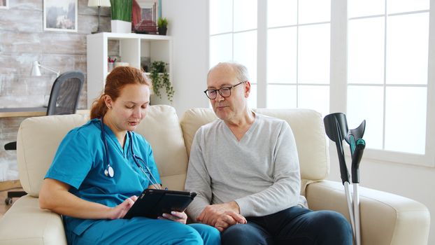 Female caucasian nurse taking notes about elderly man health in bright and cozy nursing home. Caregiver and social worker