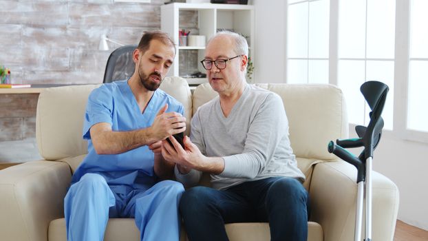 Male nurse helps a retired old man to use a smartphone in bright and cozy retirement home