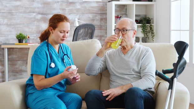 In retirement home nurse is giving daily pills to old man with crutches next to him. Caregiver and social worker