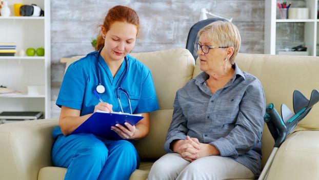 Female doctor with red hair taking notes on clipboard while talking with senior woman on the couch in nursing home