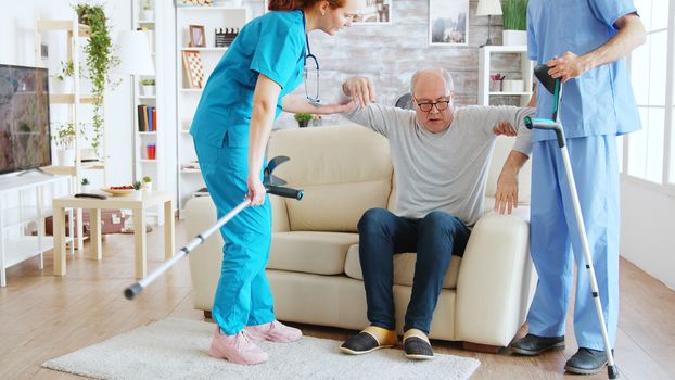 Team of nurses or social workers helping an old disabled man to walk with his crutches out of the nursing home room.