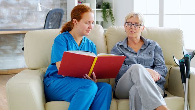 Caucasian female assistant in nursing home reading a book to an elderly retired woman who sits on the couch