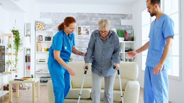 Senior woman with crutches getting help for female nurse and doctor to stand on her legs from the couch