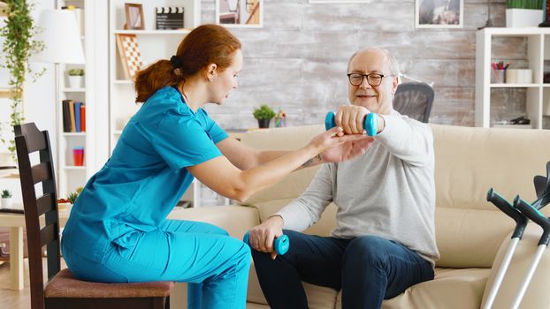 Female nurse helps an old man to make his morning exercises in bright and cozy retirement home
