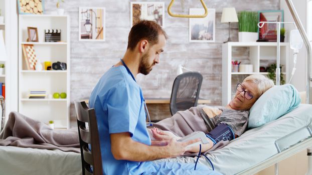 In retirement home male nurse checks old sick lady blood pressure. Nursing home worker