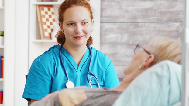 Revealing shot of female assistant talking with an old lady lying in hospital bed in a nursing home