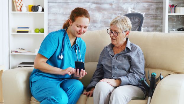 Female doctor in nursing home sitting on the couch teaching senior woman to use smartphone