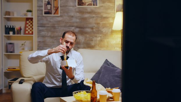 Businessman with tie eating noodles sitting on couch after a long day at work. Chinese food.