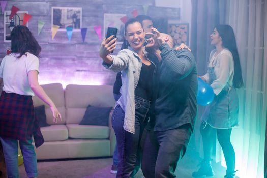 Happy young man and woman taking a selfie at a party with their friends. Group of people dancing.