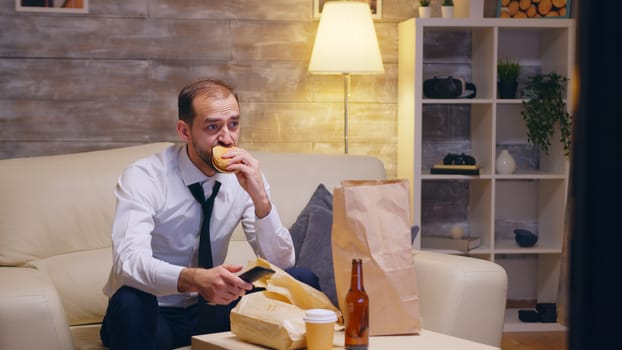 Caucasian businessman untying his tie sitting on couch after a long day at work. Eeating burger.