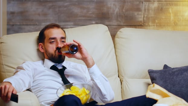 Overworked businessman sitting on couch using tv remote control and eating chips late at night.