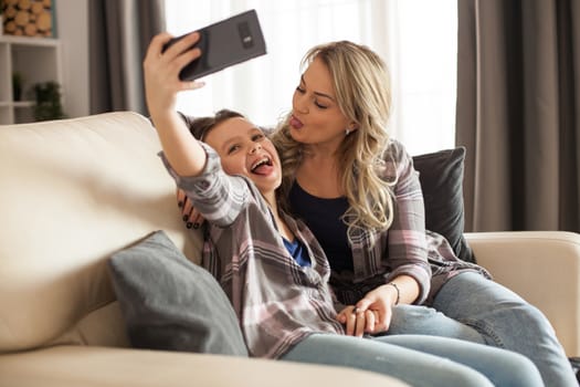 Cheerful mother and daughter sitting on the couch in living room taking a selfie together.