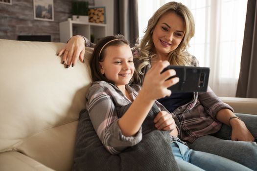 Modern mother and her little daughter watching a love video on smartphone sitting on the couch.