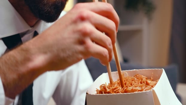 Close up of tired entrepreneur eating noodles with chopsticks in living room while watching tv.