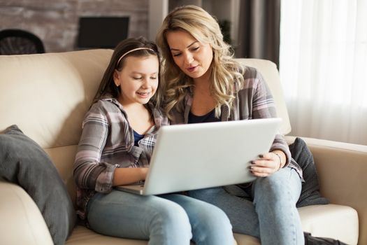 Young mother helping her little daughter to do her homework using modern laptop sitting on the couch.